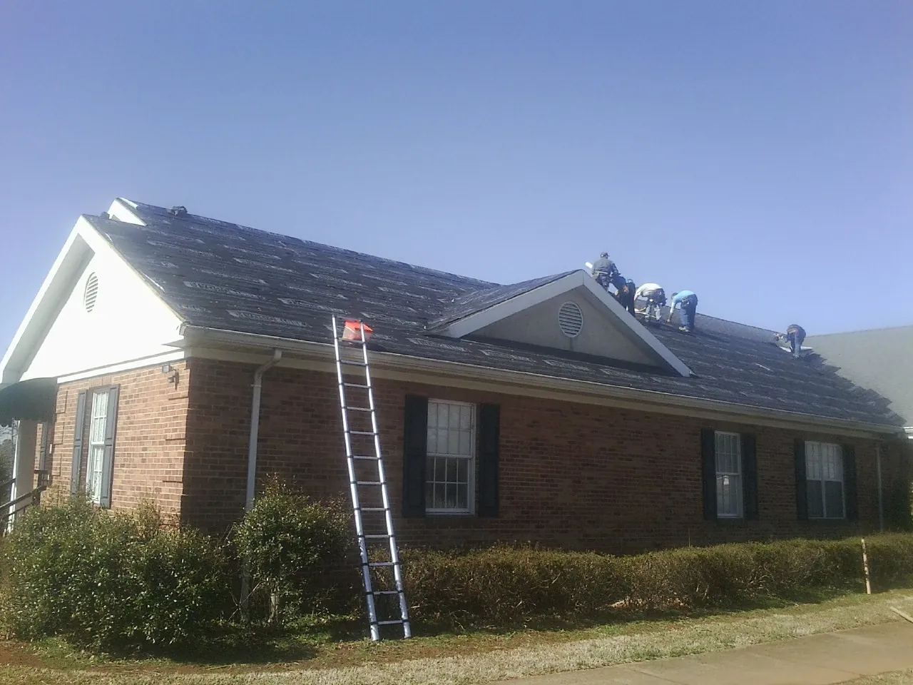Crew tearing shingles off a brick ranch roof with a ladder set — Emergency Roof Repair work by our crew in Nesbit