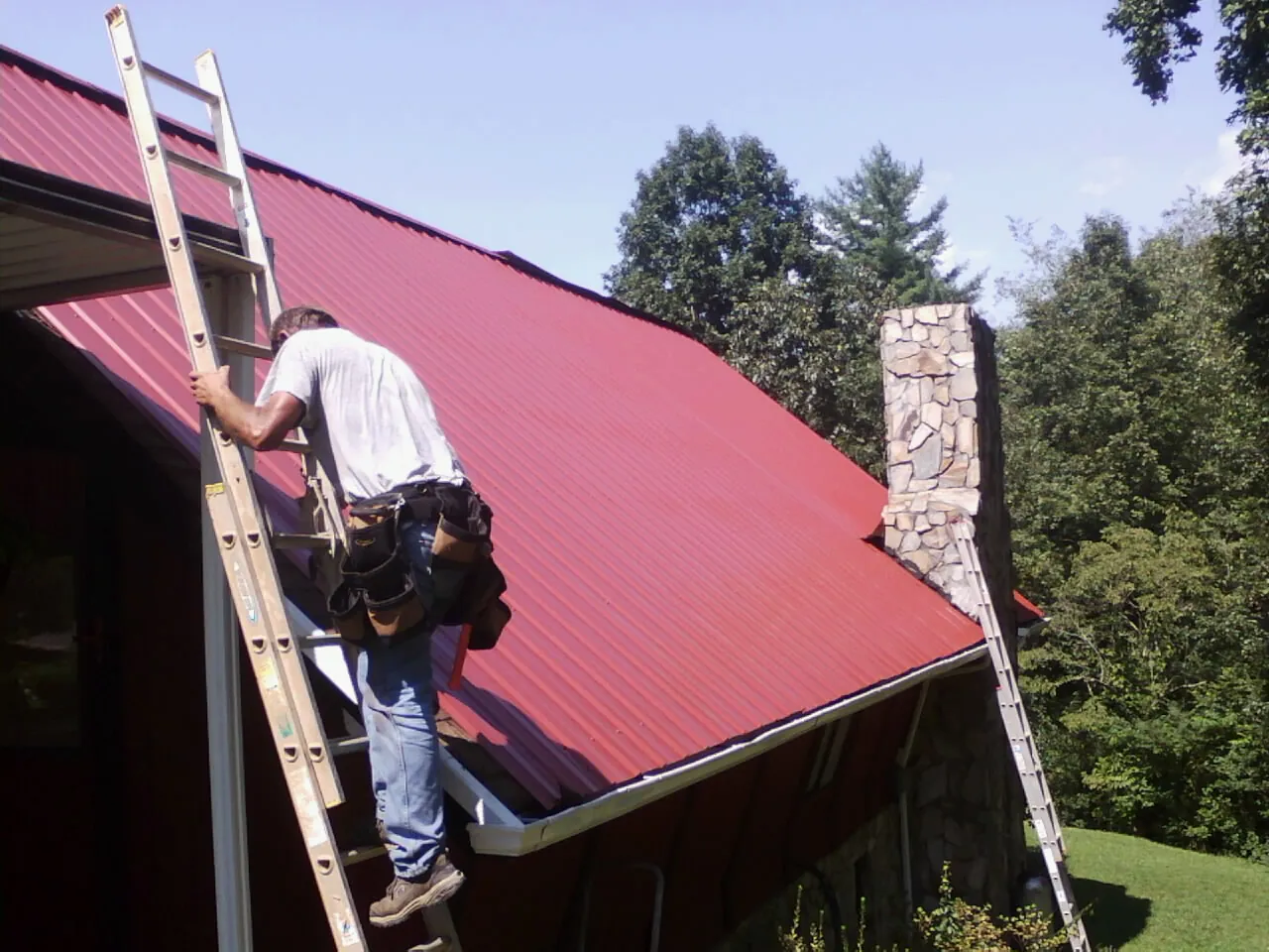 Roofer on a ladder beside a red metal roof and stone chimney — Soffit & Fascia Repair by Roofing Brothers in Nesbit